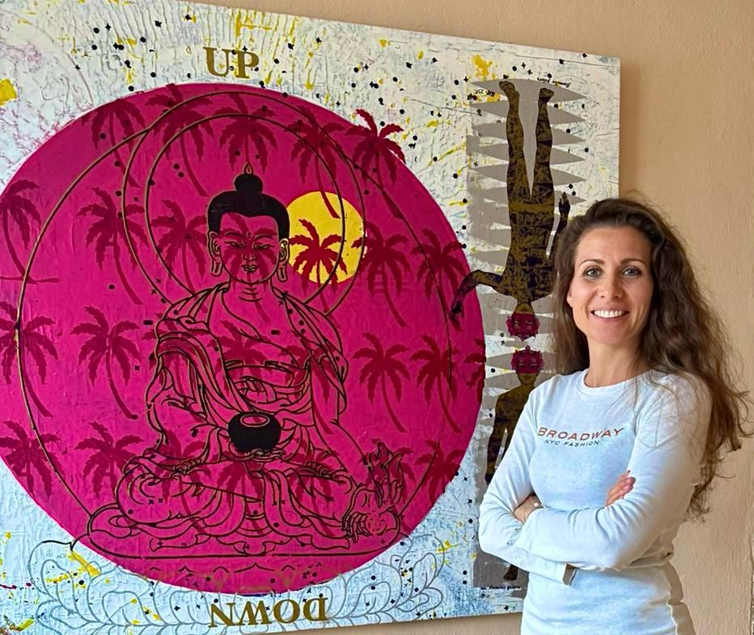 Physiotherapie Claudine König - Heilpraktikerin Physiotherapie - Eine Frau mit langen braunen Haaren lächelt neben einem großen, in Mischtechnik gefertigten Gemälde, das eine Buddha-Figur in einem rosa Kreis mit Palmen zeigt.
