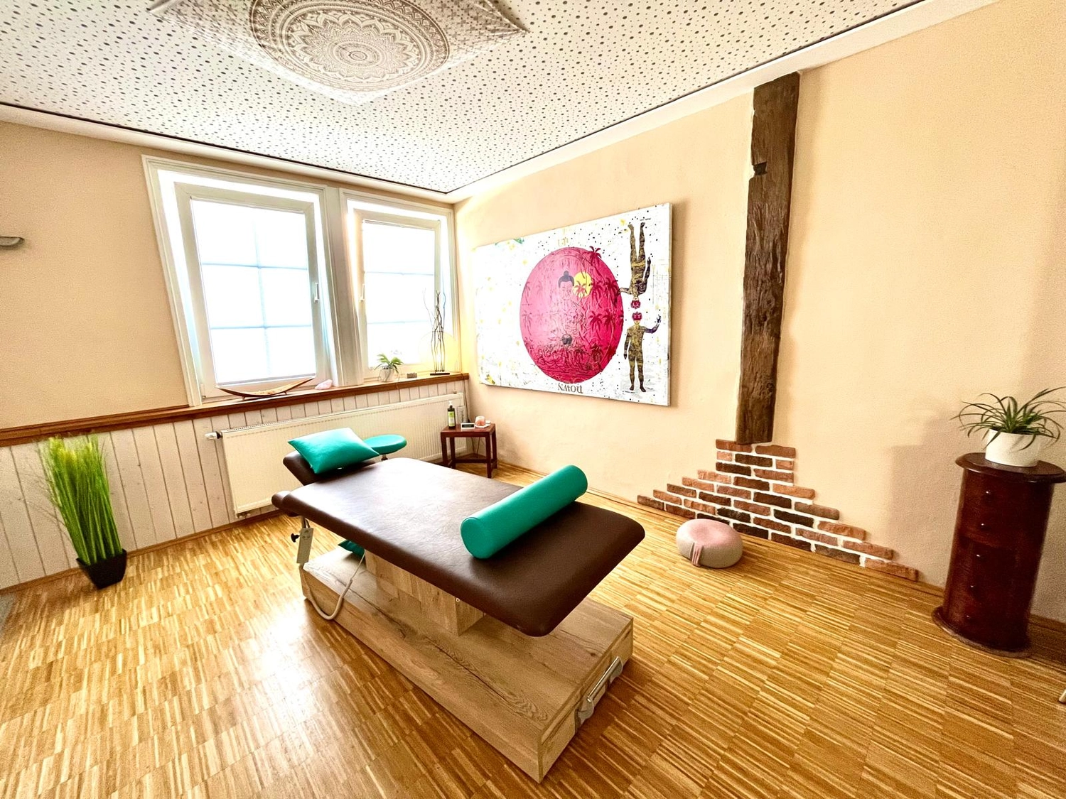 Physiotherapie Claudine König - Heilpraktikerin Physiotherapie - Ein heller Behandlungsraum mit Massageliege, Parkettboden und einem großen modernen Wandbild.
